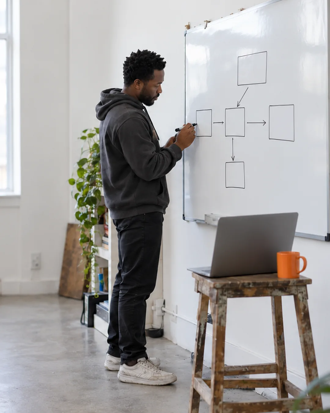 A solo founder sketches a product on a whiteboard of loose boxes and arrows in a bright co-working corner.