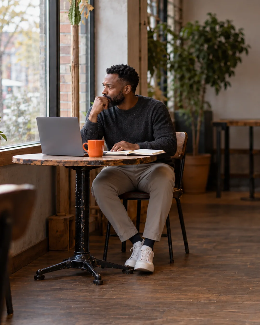 An interior designer sits by a cafe window with a laptop and notebook, thinking after a discovery call.