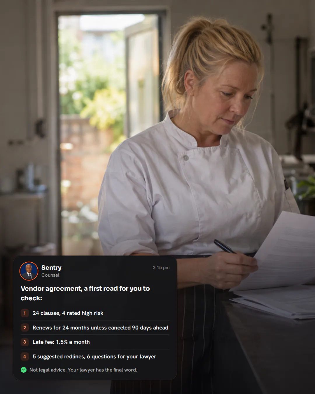 A catering company owner in chef’s whites reading a printed agreement at a steel prep table in her kitchen.