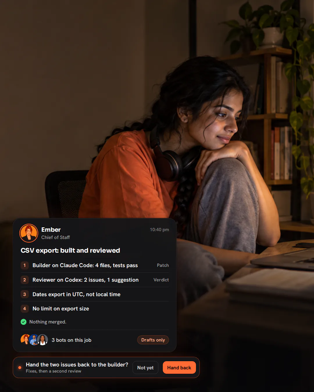 A young developer sitting cross-legged on a desk chair at night, headphones around her neck, looking at her laptop.
