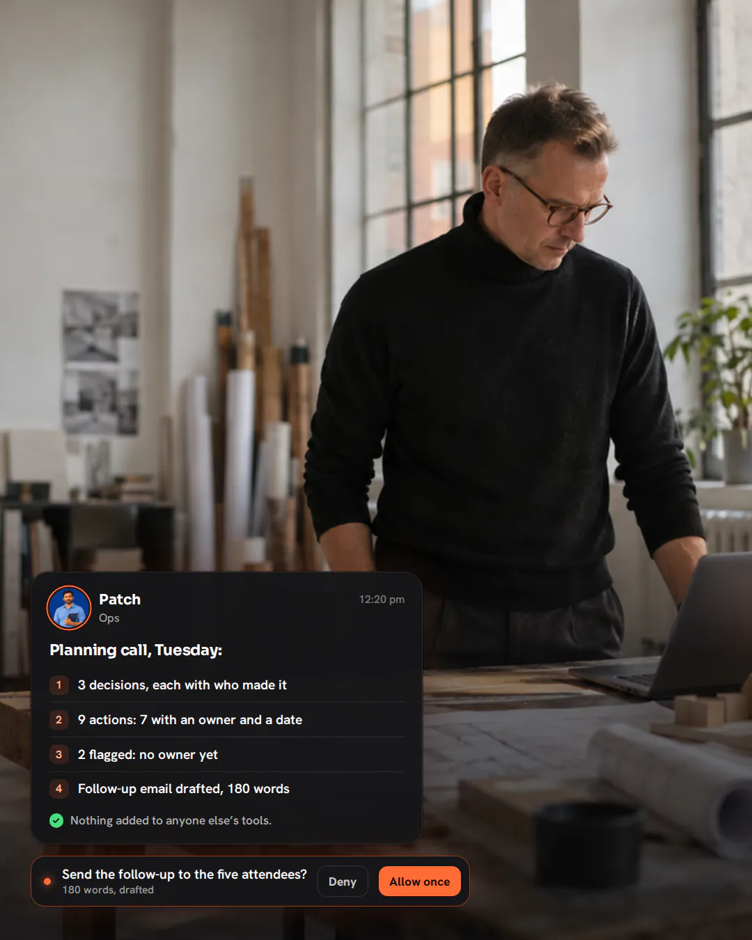 An architect in a black turtleneck standing at a drafting table in his studio, looking down at a laptop.