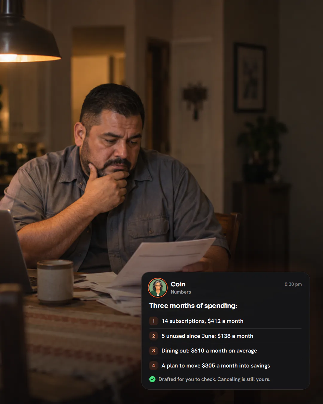 A father sitting at his kitchen table in the evening with card statements and a laptop, rubbing his jaw in thought.