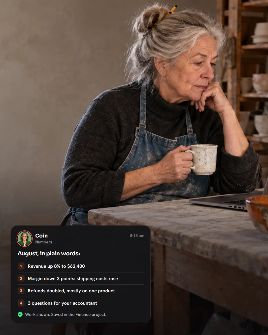 An older ceramicist in a clay-smudged apron sitting at her studio worktable with coffee, looking at a laptop beside glazed bowls.