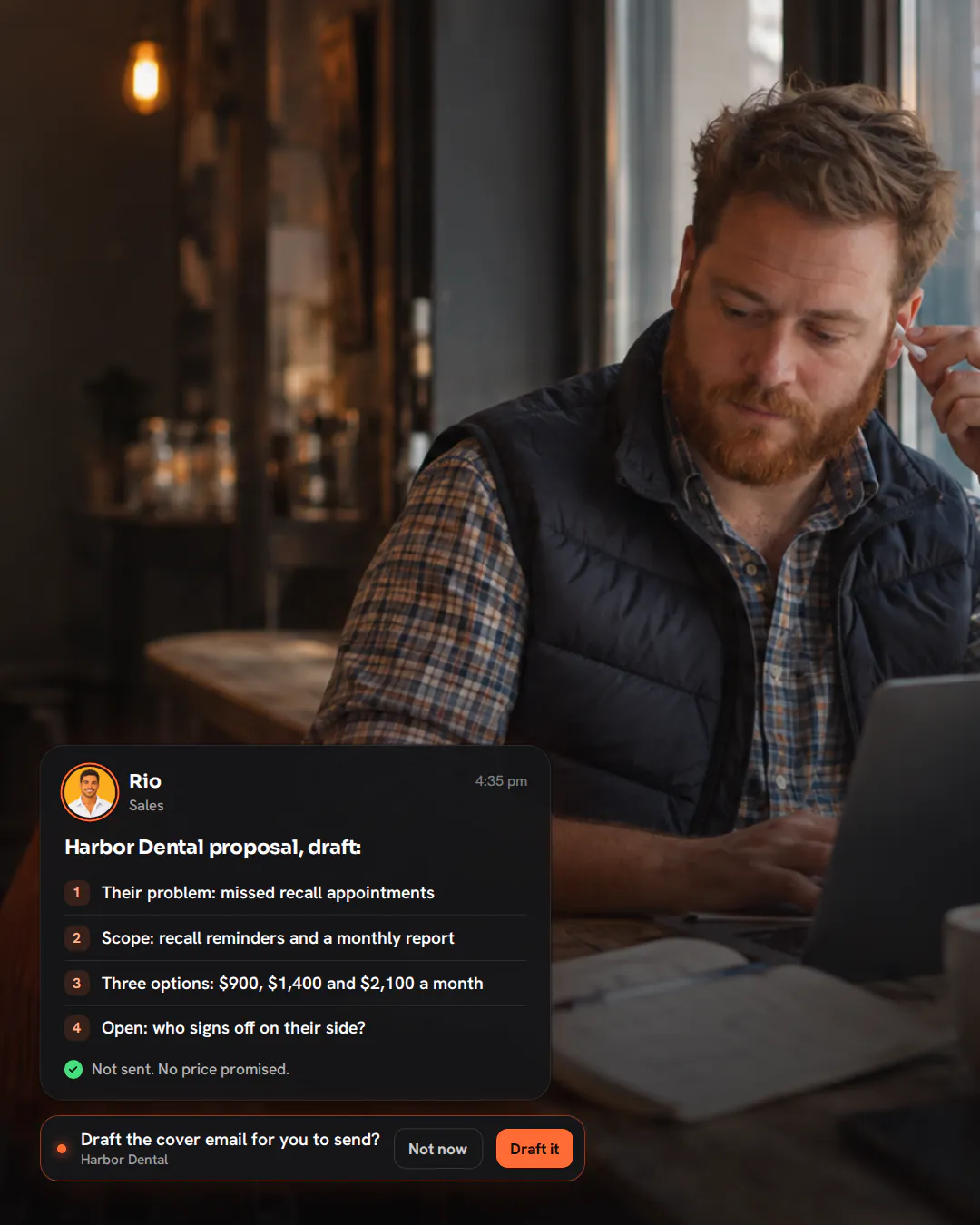 An agency owner with a ginger beard at a café window bar, taking out an earbud beside his laptop and notebook.