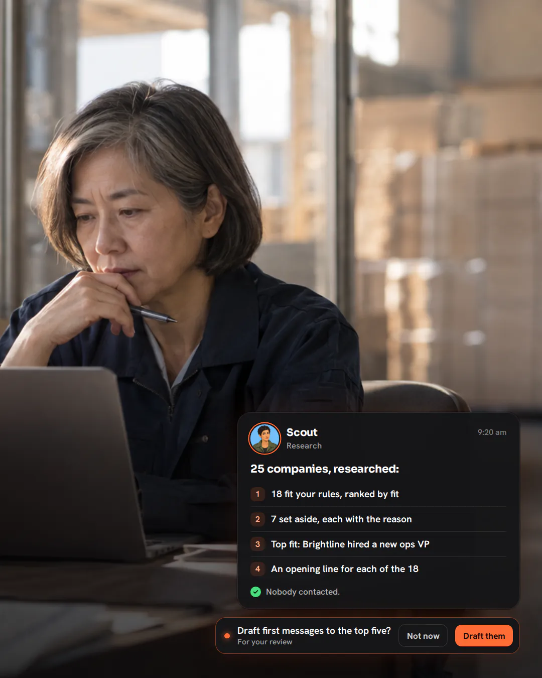 A packaging company owner at a corner desk in her warehouse office, reading a laptop with a pen in hand.